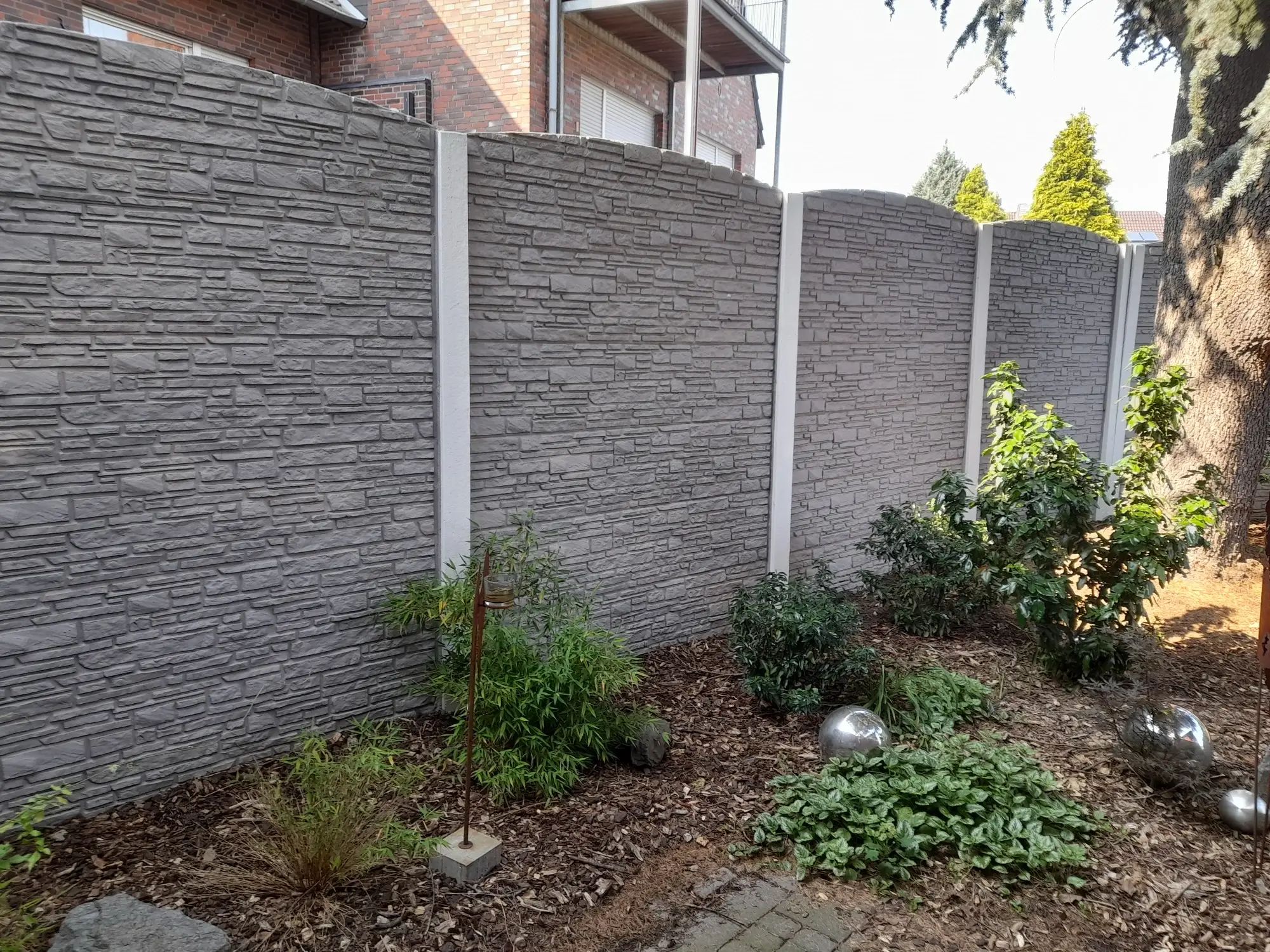 Grauer Betonzaun in Natursteinoptik mit frisch bepflanztem Beet und Rindenmulch