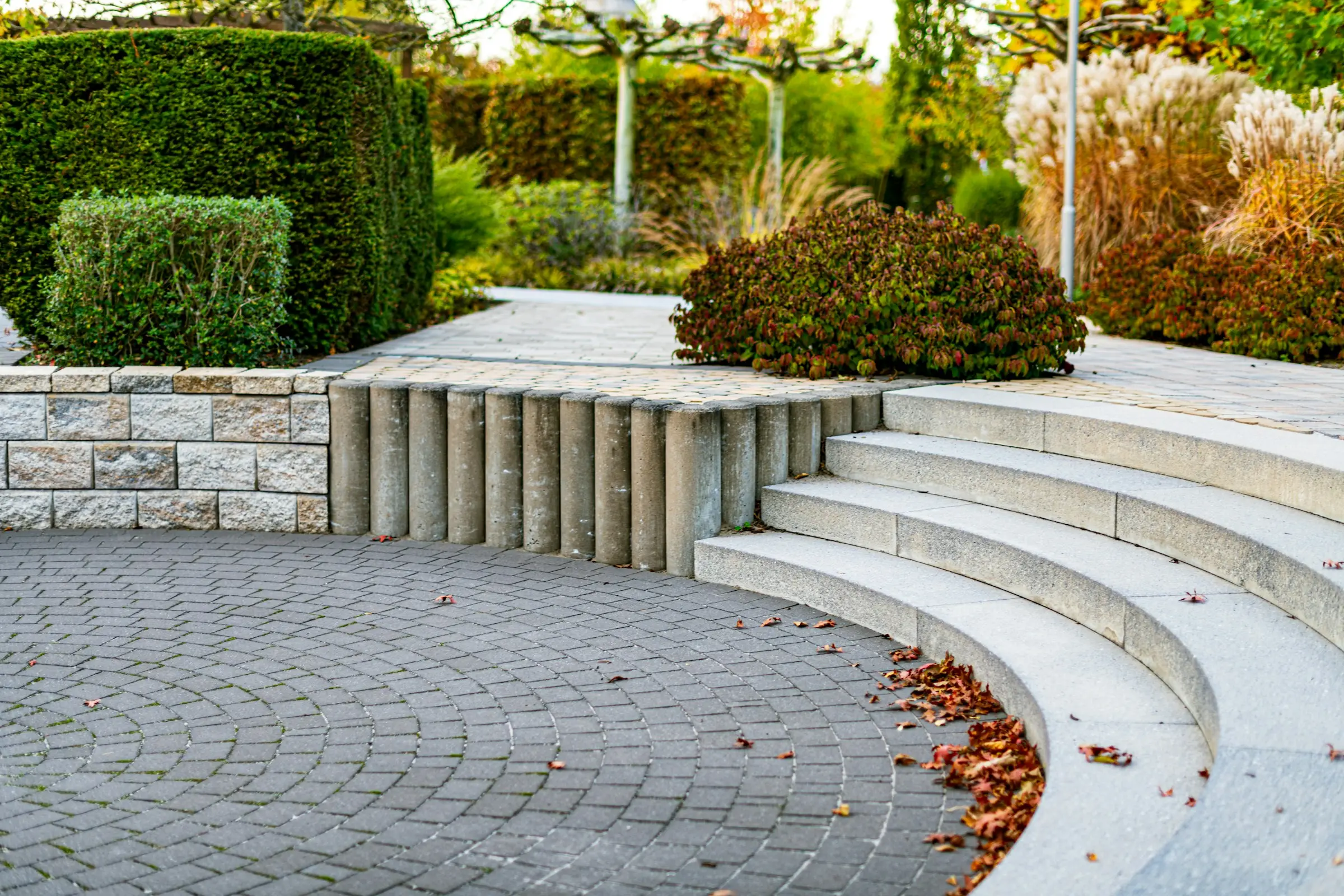 Symbolbild: gepflasterter Platz mit geschwungener Steintreppe, Natursteinmauer, Hecken und Ziergräsern